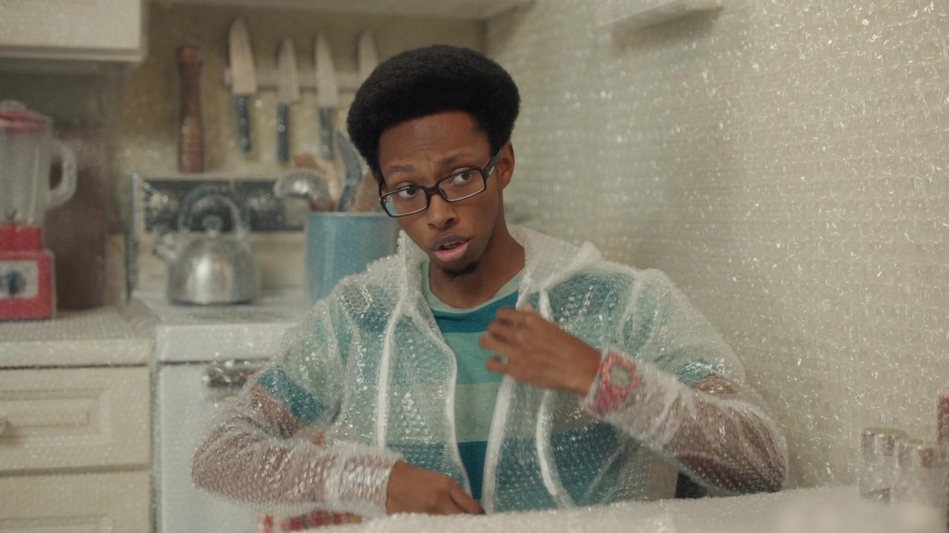 A man wears a bubble-wrap hoodie in a kitchen lined with bubble wrap.